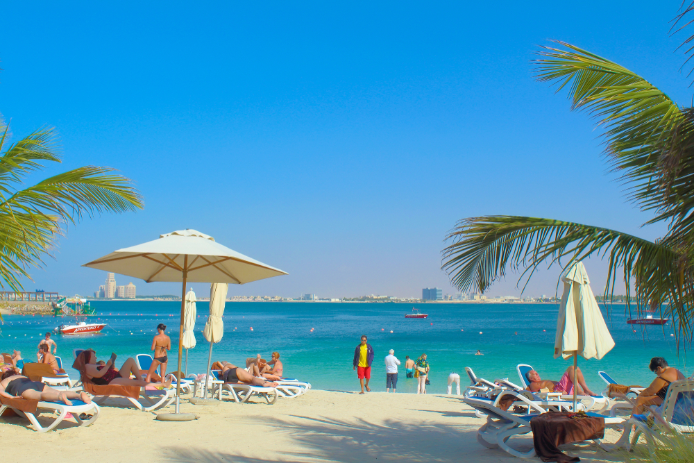 White sand beach with loungers, cream umbrellas, palm trees, and turquoise Arabian Gulf waters at Al Marjan Island beach club lifestyle destination