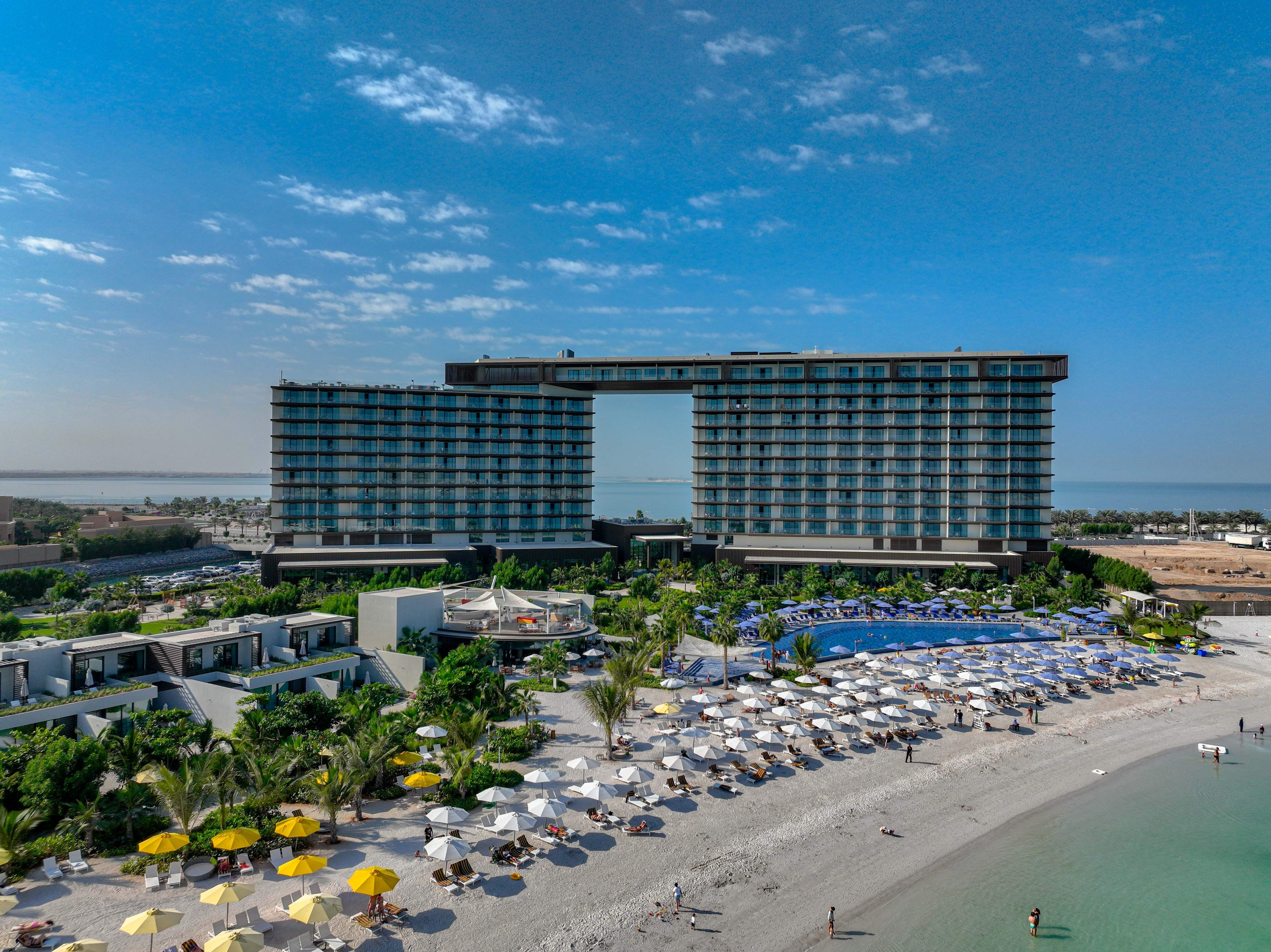Aerial view of luxury beachfront resort with twin towers connected by sky bridge, expansive pool complex, white sand beach, and turquoise waters at Al Marjan Island